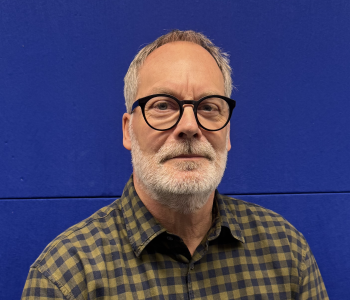 Mann vor blauem Hintergrund. Trägt Brille und ein kariertes Hemd.